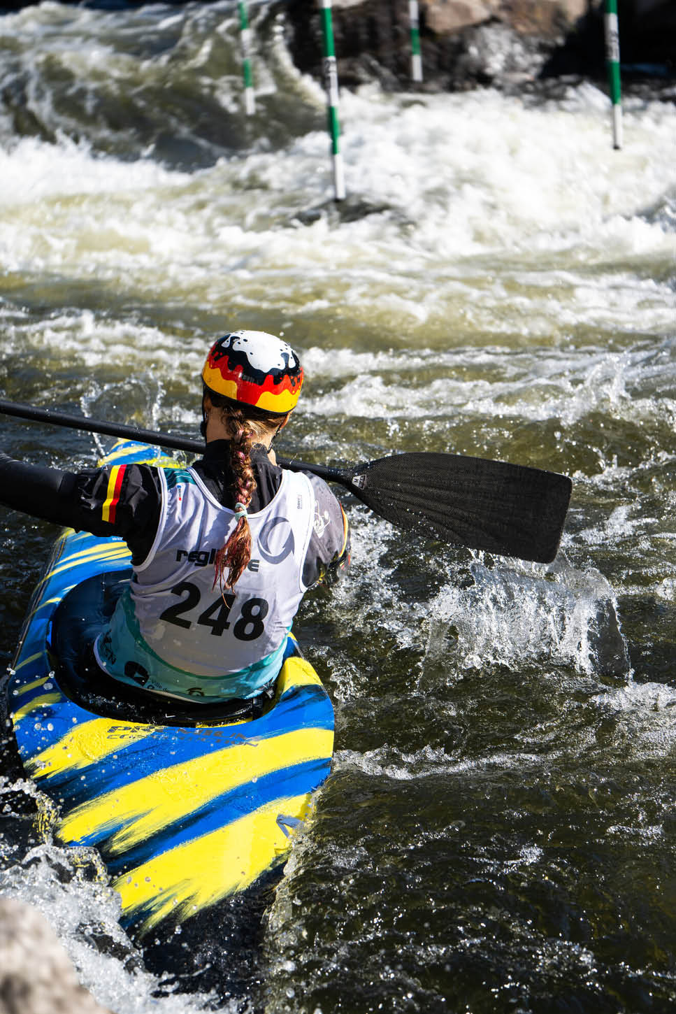 Eine Person mit Helm und Trikot paddelt in einem bunten Kajak durch wildes Wasser bei einem Kanuslalom-Wettbewerb. Beschreibung von KI generiert