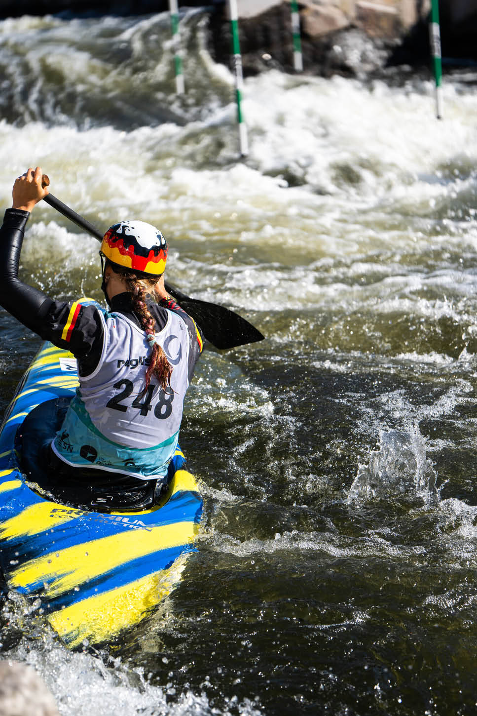Eine Person mit Helm und Trikot paddelt in einem gelb-blauen Kajak durch wildes Wasser bei einem Kanuslalom-Wettbewerb. Beschreibung von KI generiert