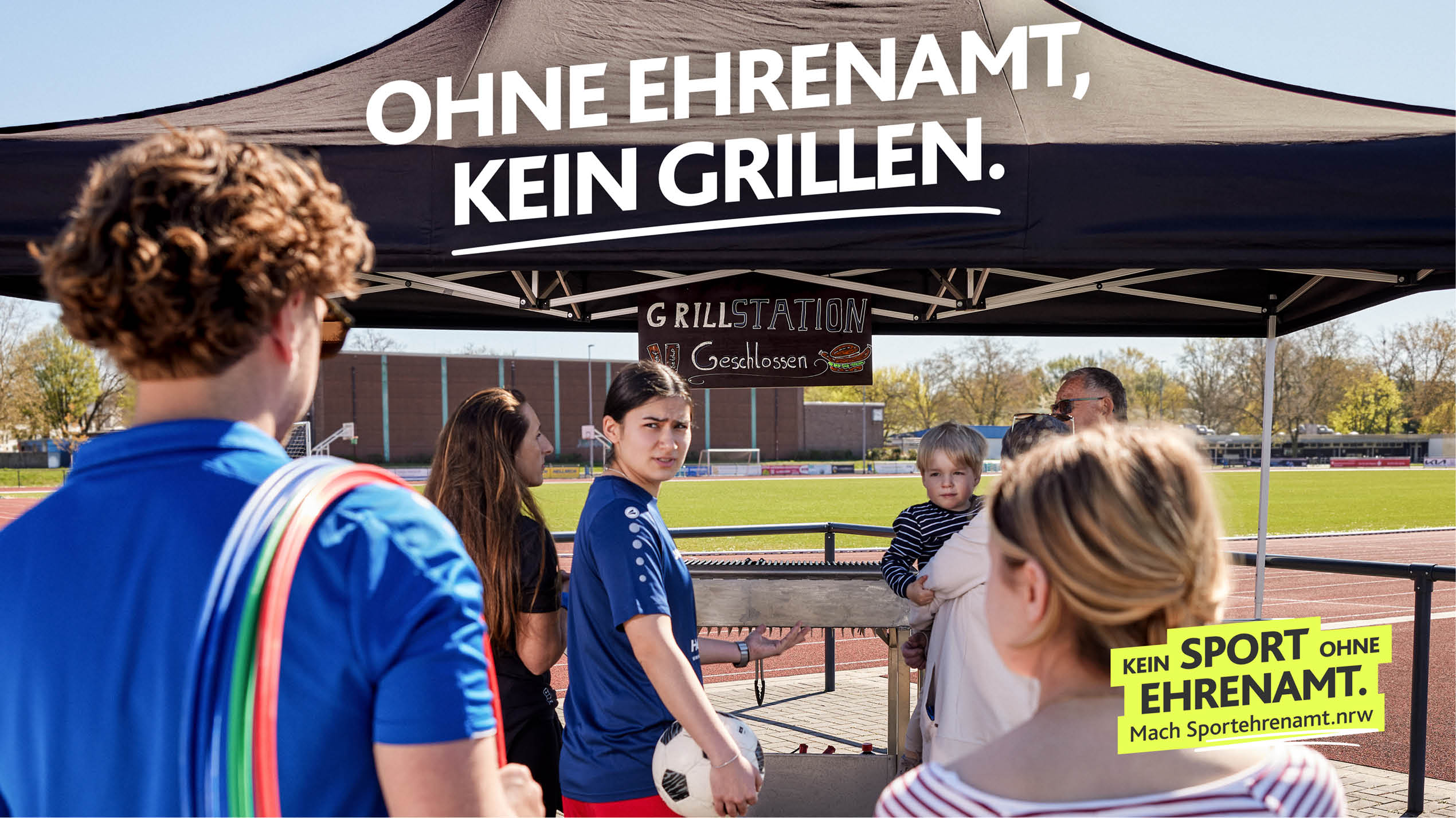 Mehrere Personen stehen unter einem Zelt mit der Aufschrift „Ohne Ehrenamt, kein Grillen“ und einem Schild, das f r Sport ohne Ehrenamt wirbt, auf einem Sportplatz. Beschreibung von KI generiert