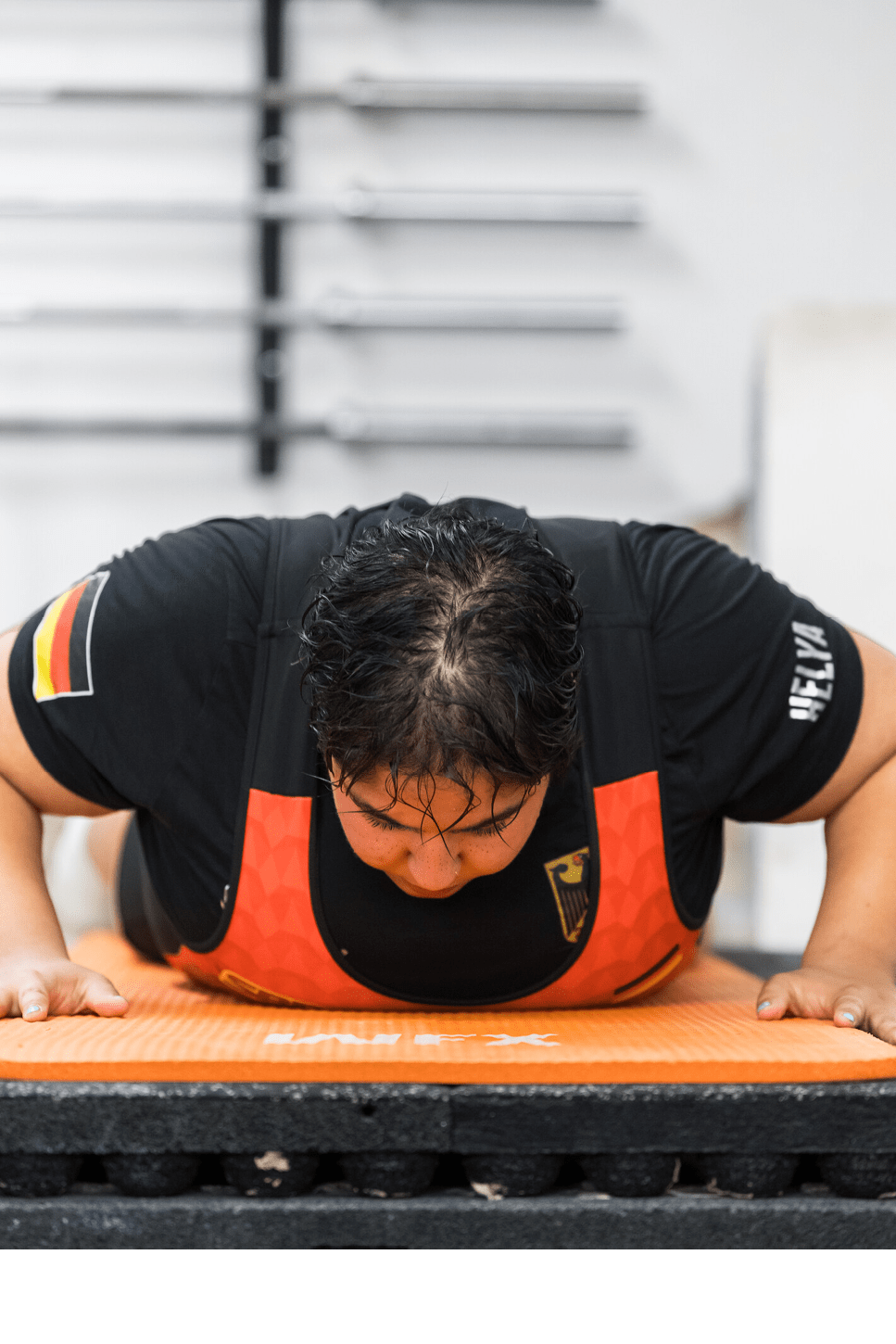 Eine Person mit schwarzem T-Shirt und rotem Trainingsanzug macht Liegest tze auf einer orangen Matte in einem Fitnessstudio. Beschreibung von KI generiert