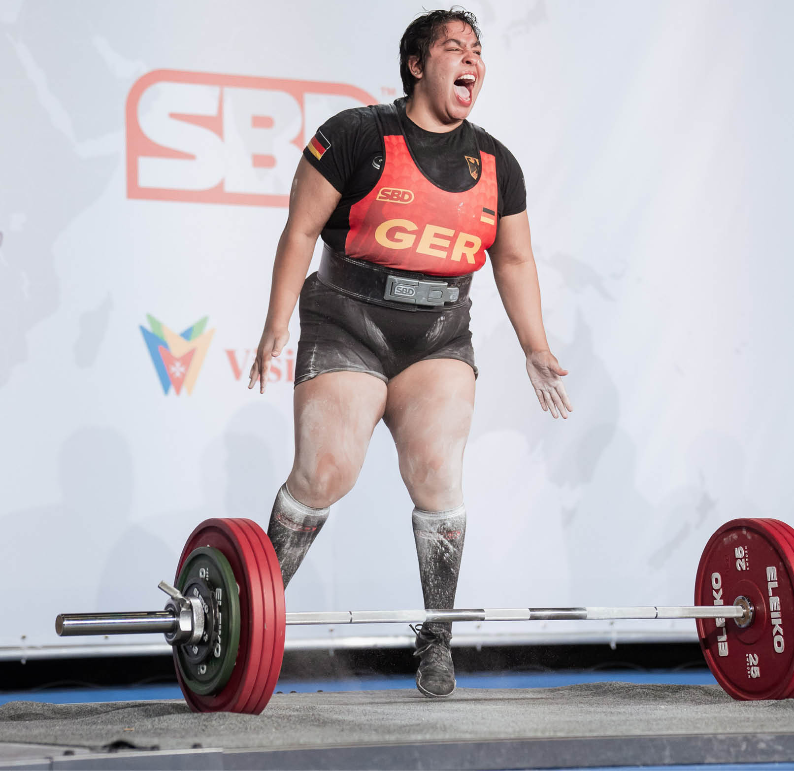 Eine Person in Sportkleidung mit der Aufschrift GER zeigt eine kraftvolle Emotion beim Gewichtheben auf einer B hne mit Sponsorenlogos im Hintergrund. Beschreibung von KI generiert