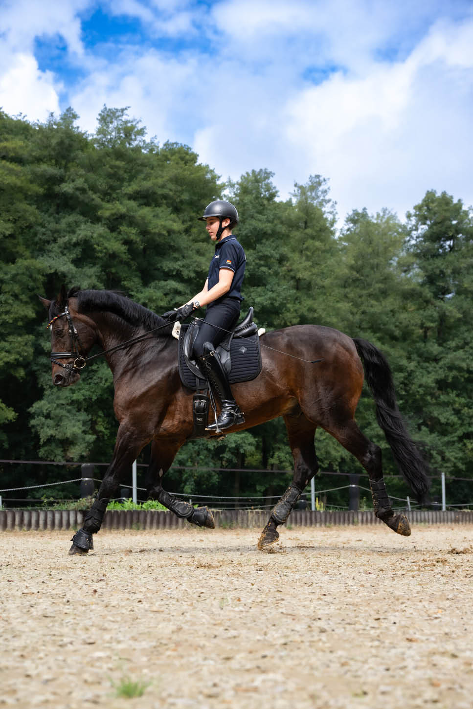 Eine Person in Reitkleidung und Helm reitet auf einem braunen Pferd auf einem Sandplatz vor einem Hintergrund aus B umen und blauem Himmel. Beschreibung von KI generiert