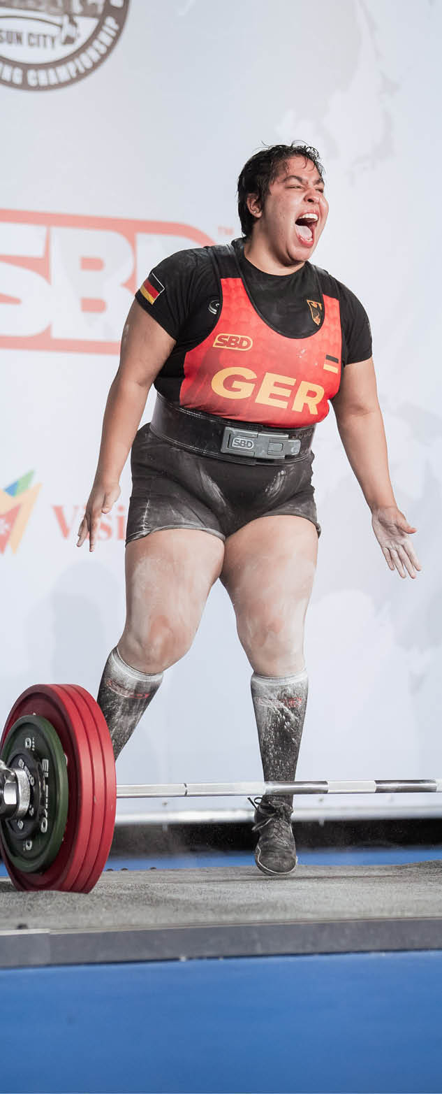 Eine Person in Sportkleidung mit der Aufschrift GER zeigt eine kraftvolle Emotion beim Gewichtheben auf einer B hne mit Sponsorenlogos im Hintergrund. Beschreibung von KI generiert