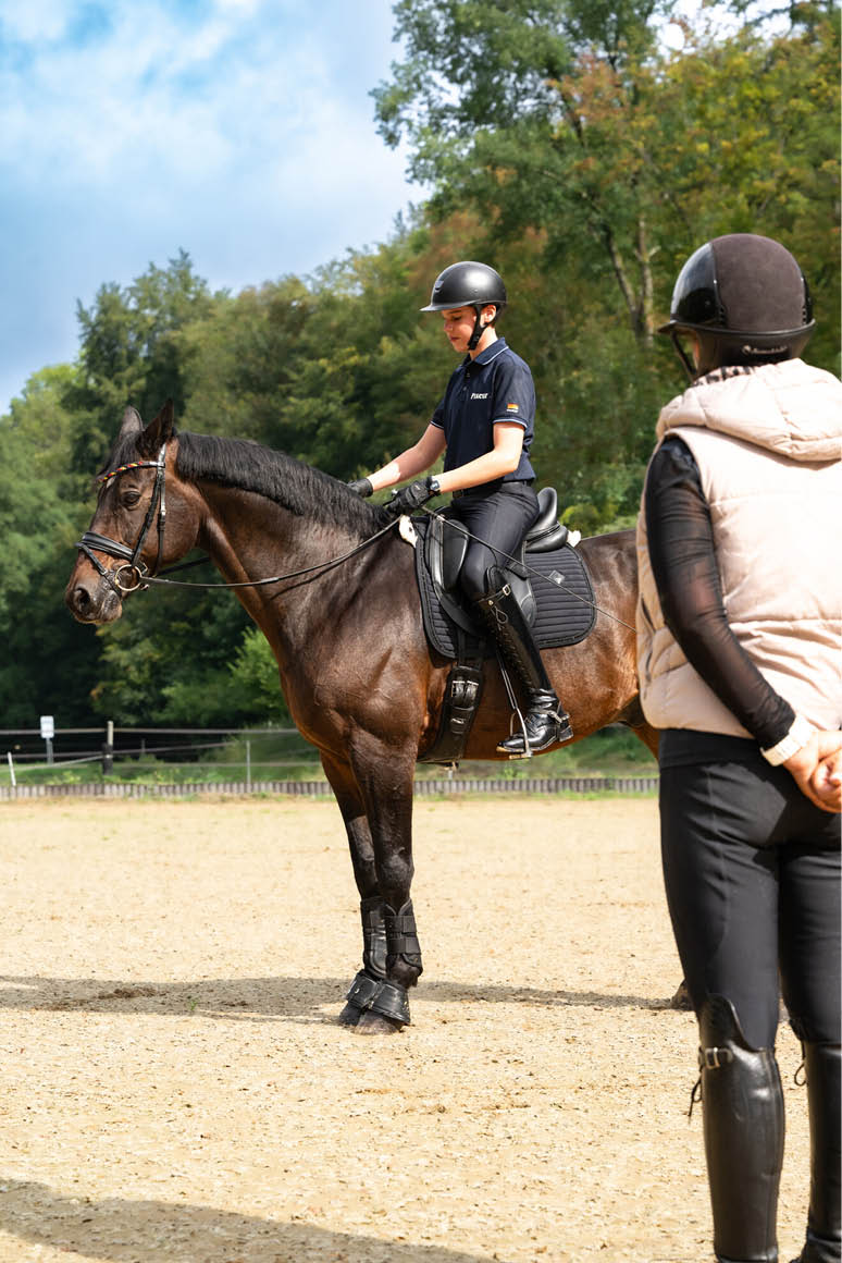 Eine Person in Reitkleidung sitzt auf einem braunen Pferd auf einem Reitplatz, w hrend eine weitere Person mit Helm und Weste daneben steht und zuschaut. Beschreibung von KI generiert