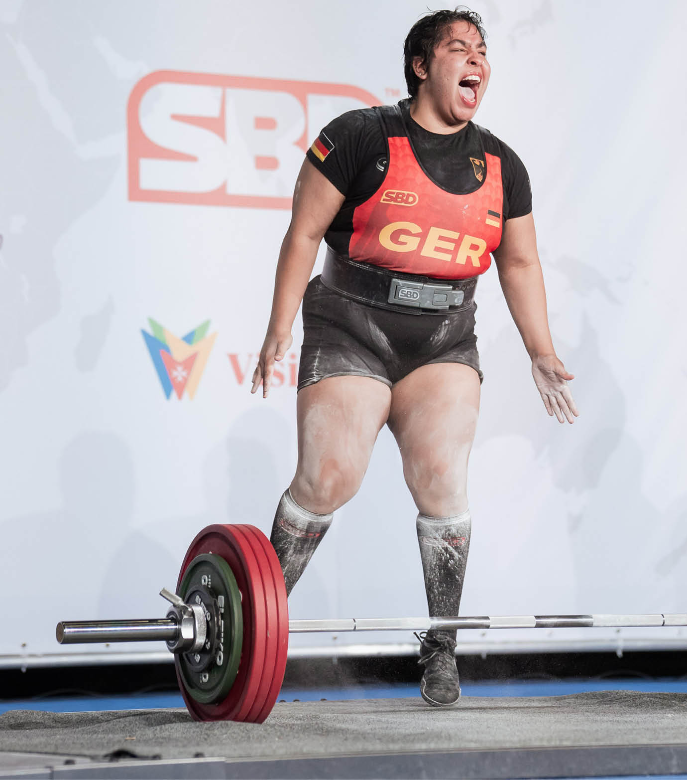 Eine Person in Sportkleidung mit der Aufschrift GER zeigt eine kraftvolle Emotion beim Gewichtheben auf einer B hne mit Sponsorenlogos im Hintergrund. Beschreibung von KI generiert