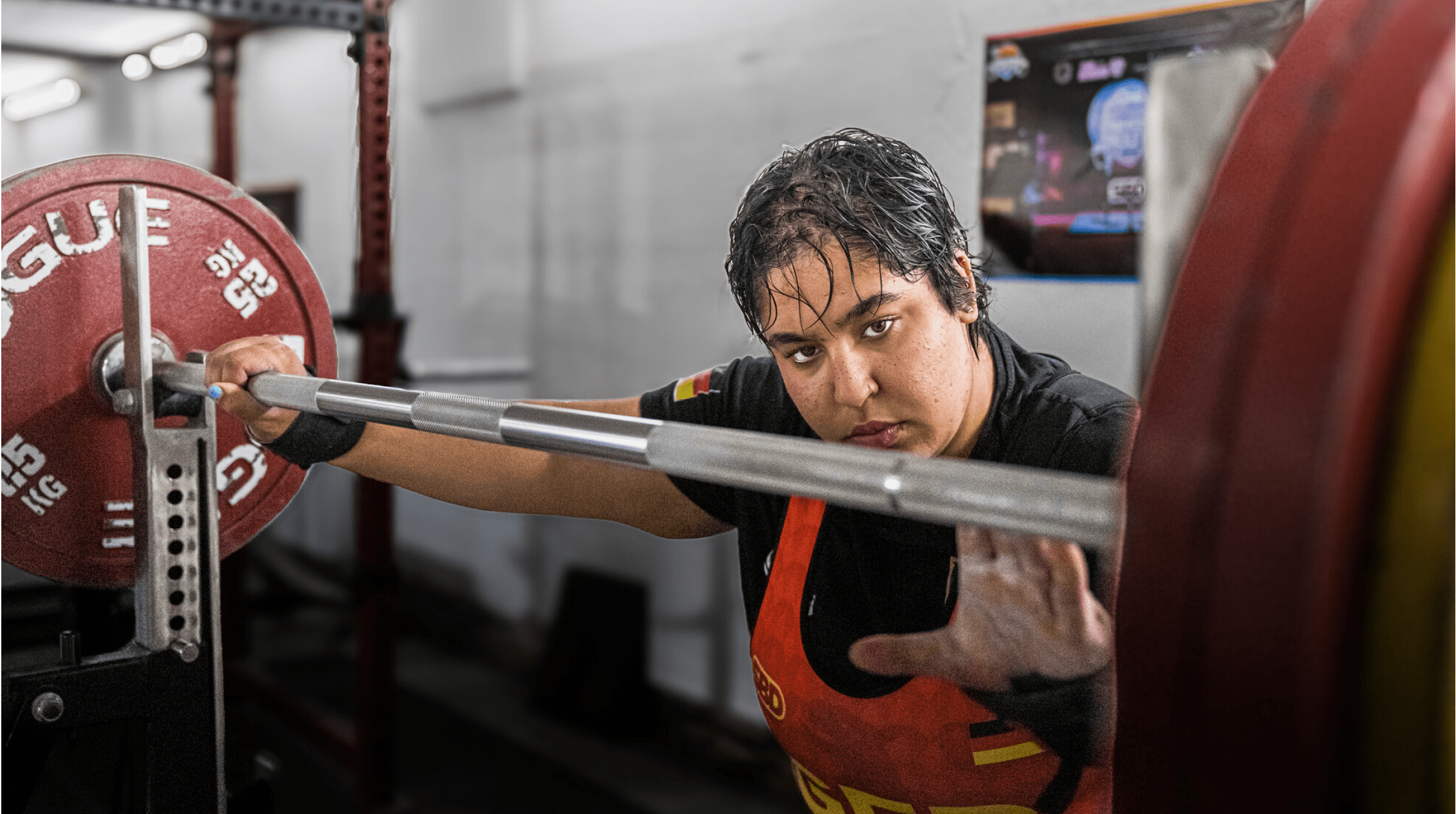 Eine Person mit nassem Haar und rotem Trainingsanzug bereitet sich in einem Fitnessstudio darauf vor, eine schwere Langhantel mit Gewichten zu heben. Beschreibung von KI generiert
