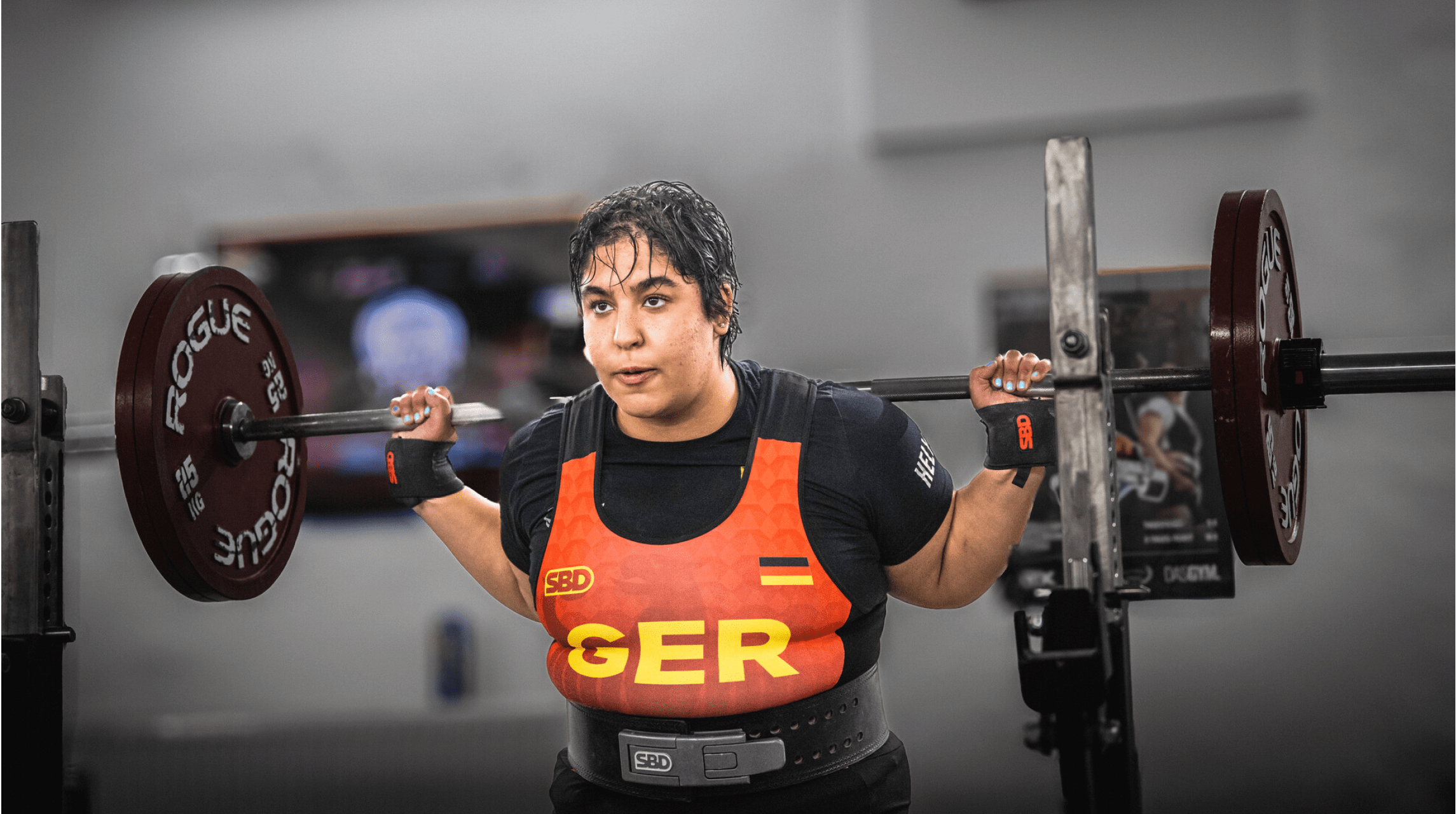 Eine Person mit nassen Haaren tr gt ein Gewichtheber-Trikot mit der Aufschrift GER und hebt eine Langhantel in einem Fitnessstudio. Beschreibung von KI generiert