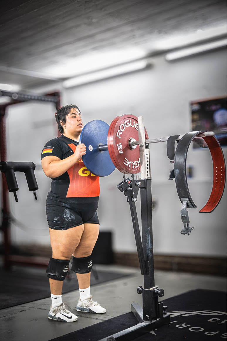 Eine Person mit kurzen dunklen Haaren tr gt Sportkleidung und Kniebandagen und steht in einem Fitnessstudio vor einer Langhantel mit Gewichten. Beschreibung von KI generiert