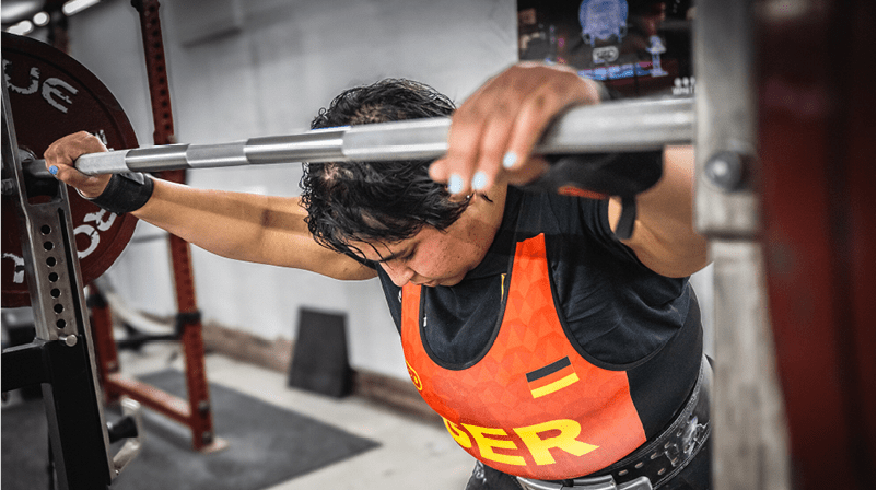 Eine Person mit kurzem dunklem Haar tr gt ein rotes Sporttrikot und hebt konzentriert eine Langhantel im Fitnessstudio. Beschreibung von KI generiert