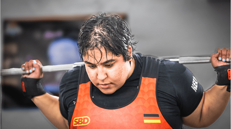 Eine Person mit kurzen nassen Haaren tr gt eine Hantelstange auf den Schultern und tr gt ein oranges Sporttrikot. Beschreibung von KI generiert